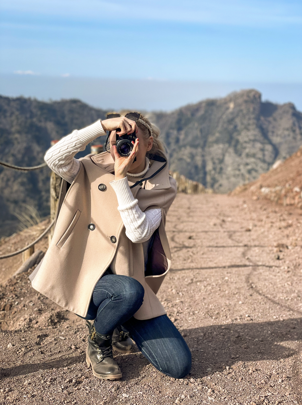 Greta Hart crouching on the trail at Mount Vesuvius, Italy, taking a photo with a film camera. Sunny day with mountains in the background. Wearing a beige cape coat, cream cable-knit sweater, jeans, and black boots.