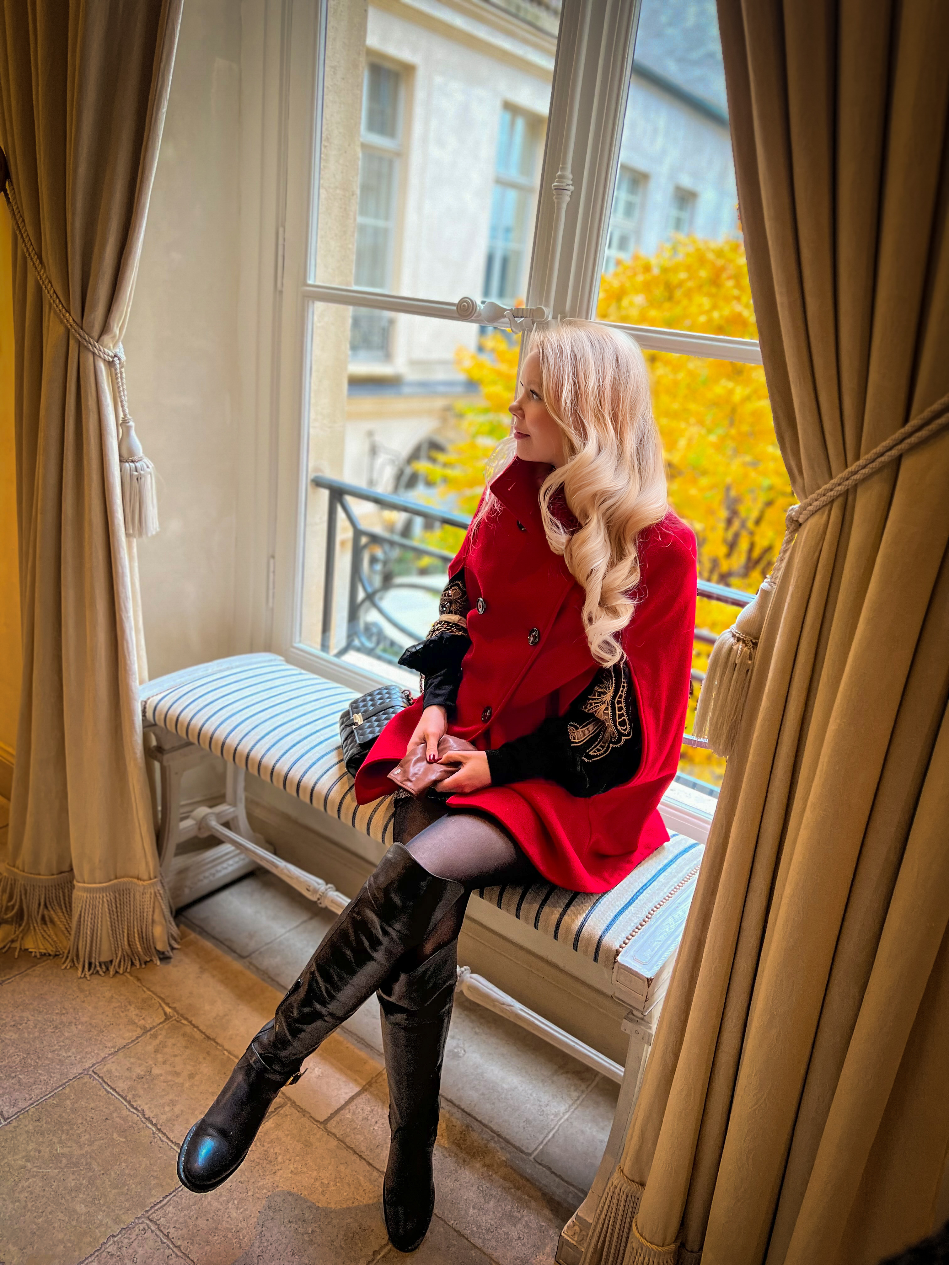 Greta Hart sitting by a window in a red coat, looking out at autumn leaves